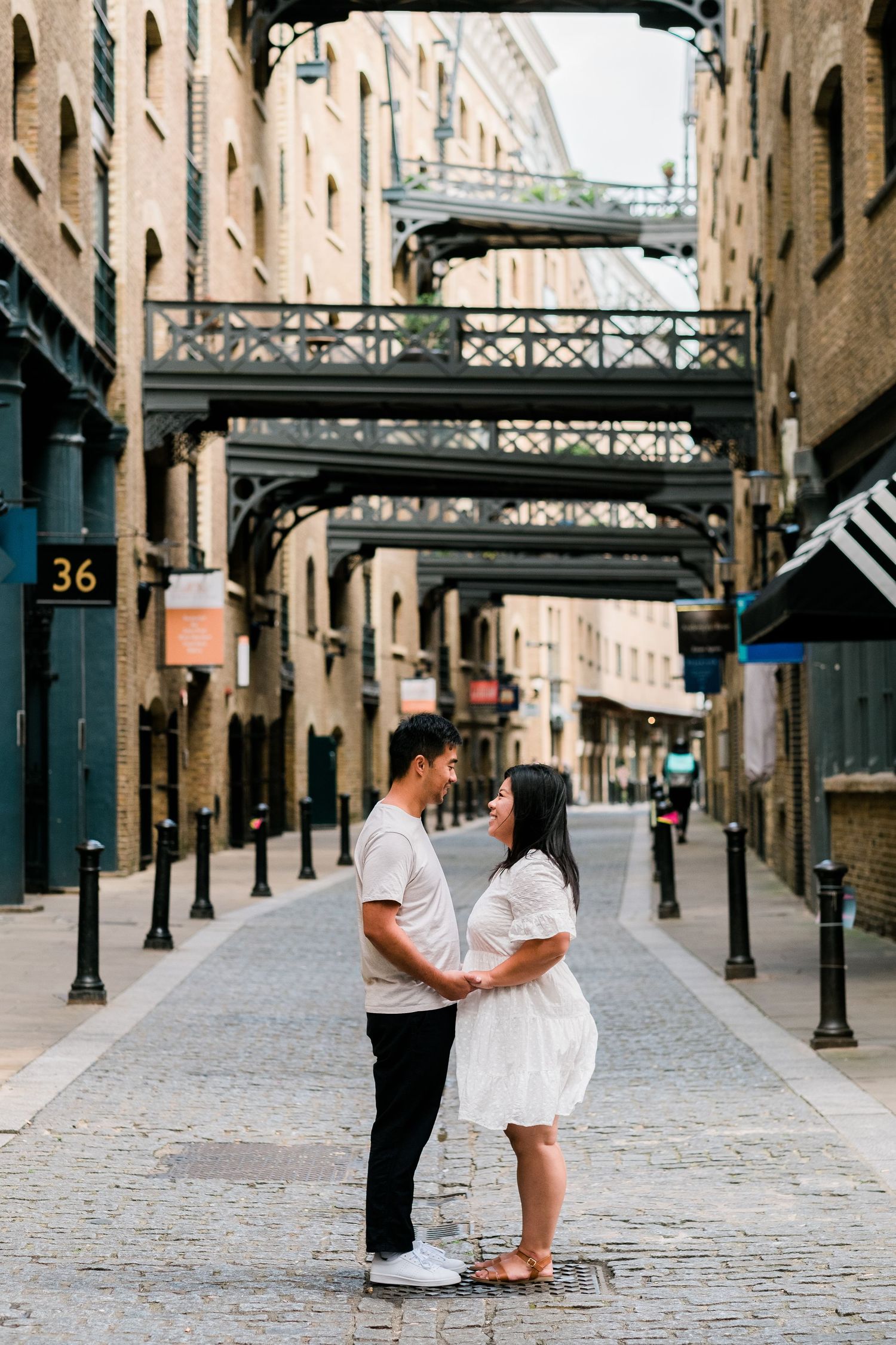 Couple embracing on cobblestone street with historic warehouse bridges overhead.