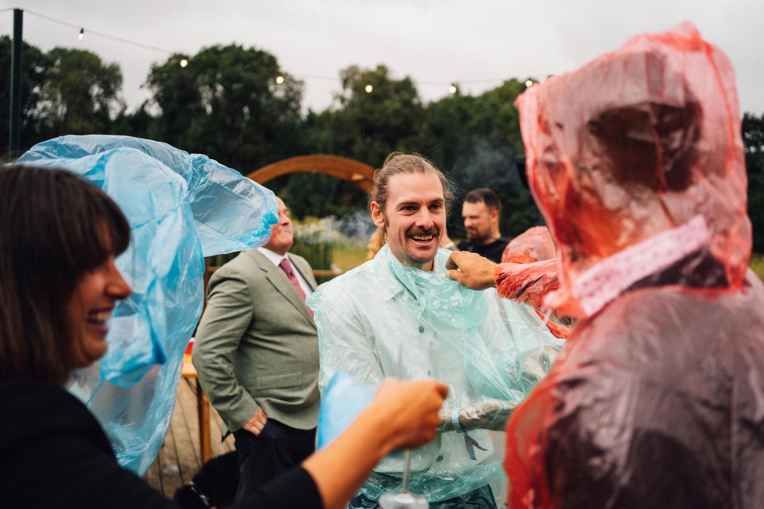 Wedding guests in rain ponchos share laughs during an outdoor celebration.