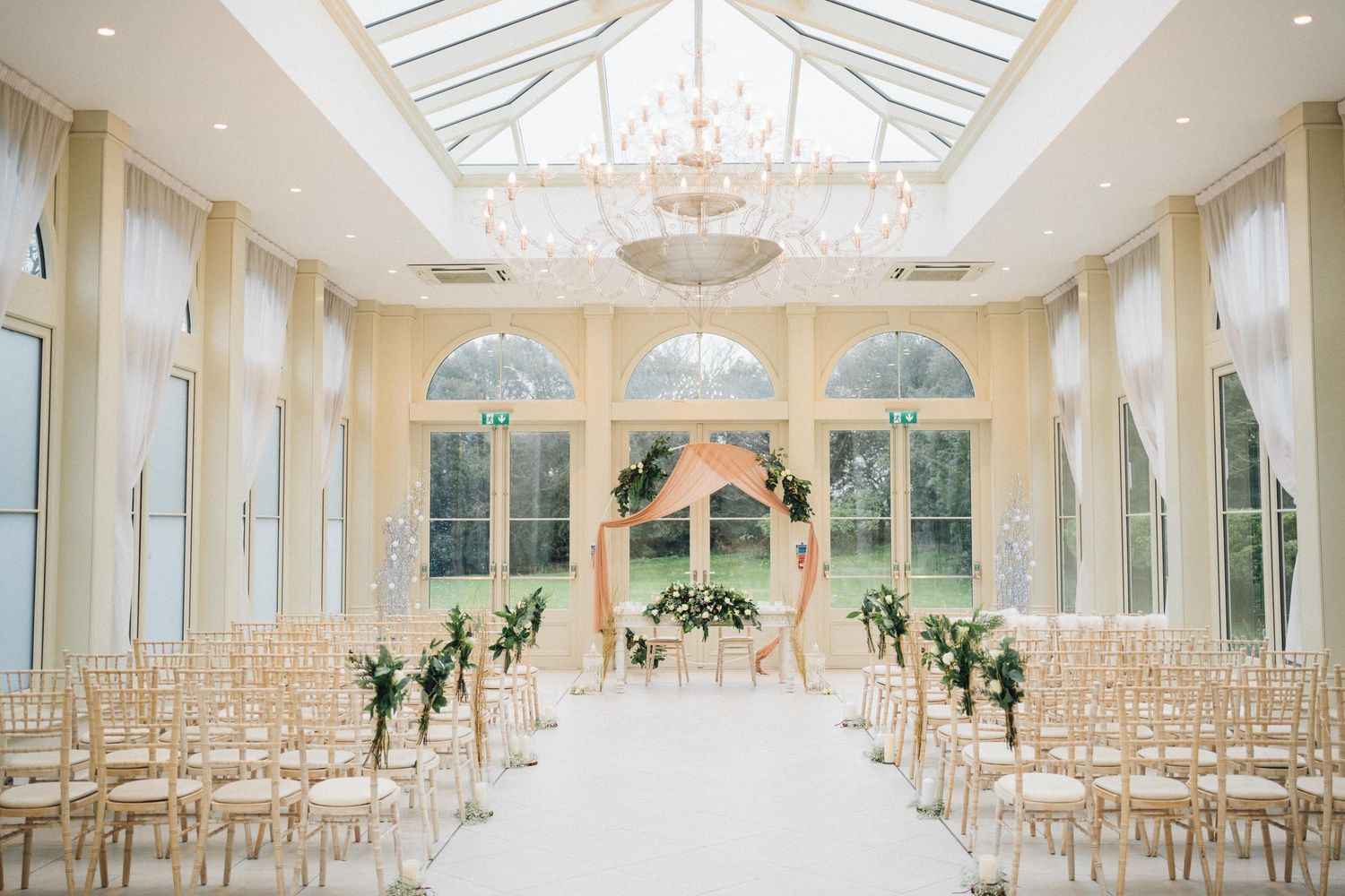 Elegant white wedding venue with glass ceiling, chandeliers and rows of chairs set for ceremony.