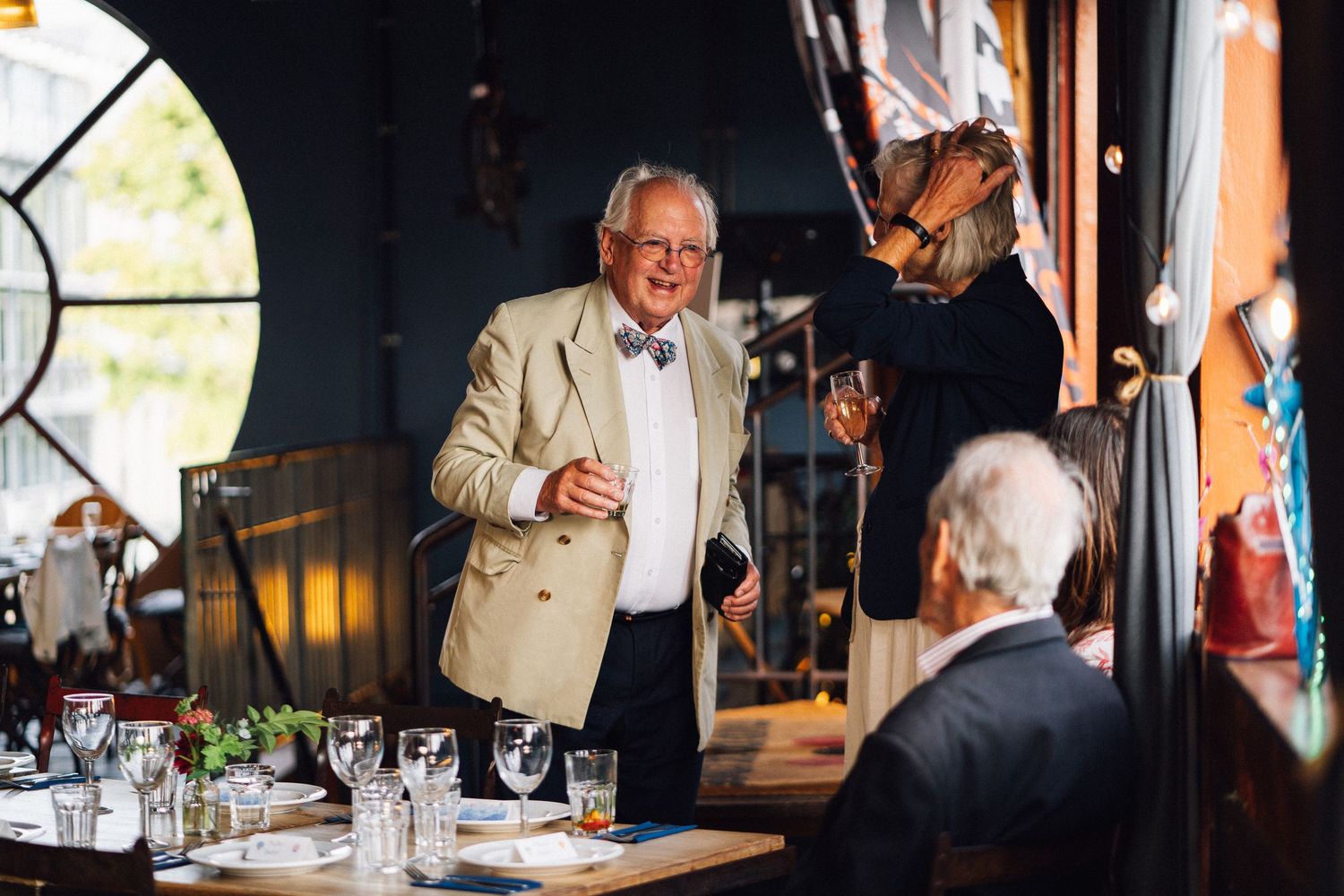 A speaker in a beige suit gestures while giving a speech at an elegant dinner event.