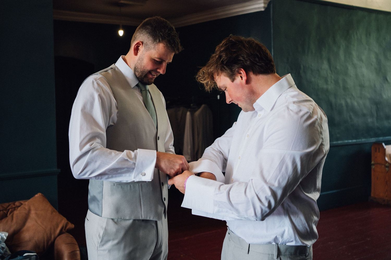 Two people in formal attire adjust their outfits while getting ready in a dimly lit room.