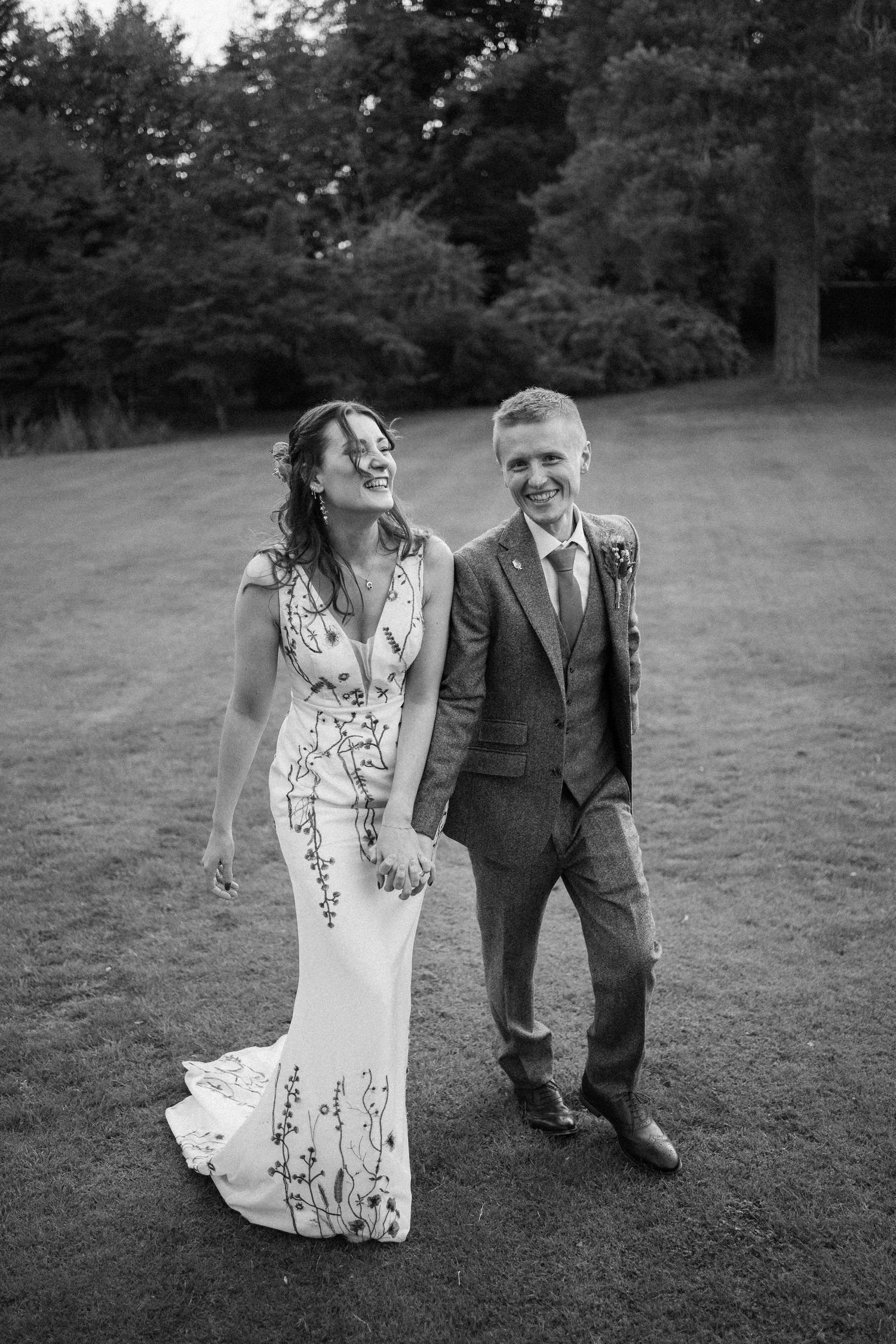 A joyful wedding couple laughs together on a grassy lawn with trees in the background, captured in black and white.