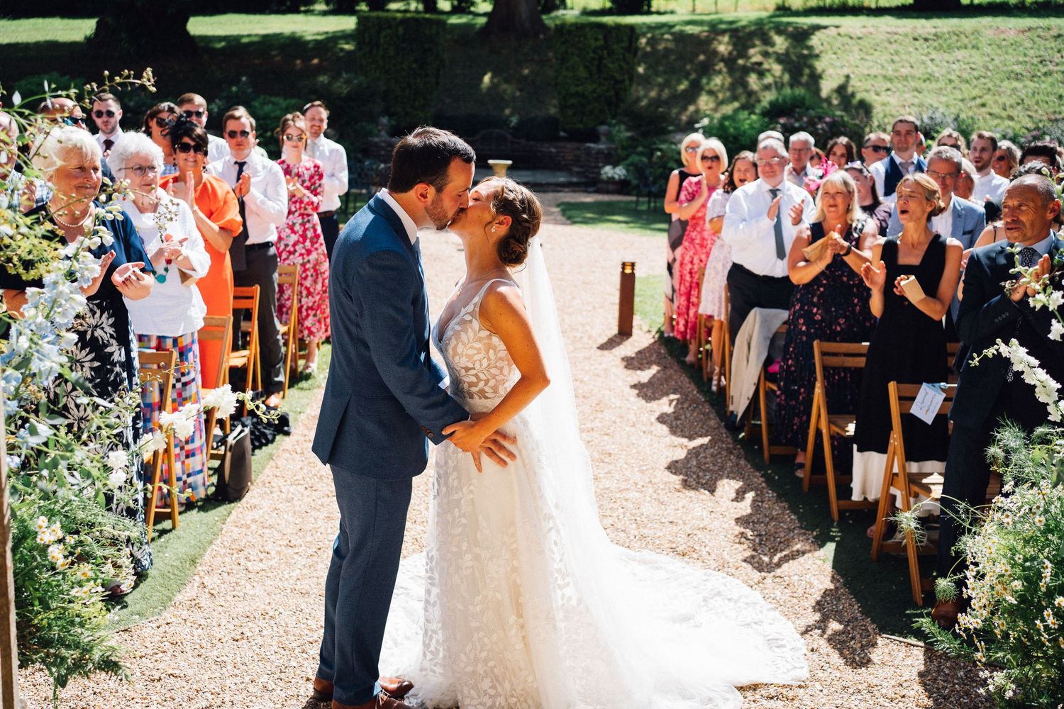 A wedding party stands outdoors on a sunny day surrounded by greenery and garden landscaping.