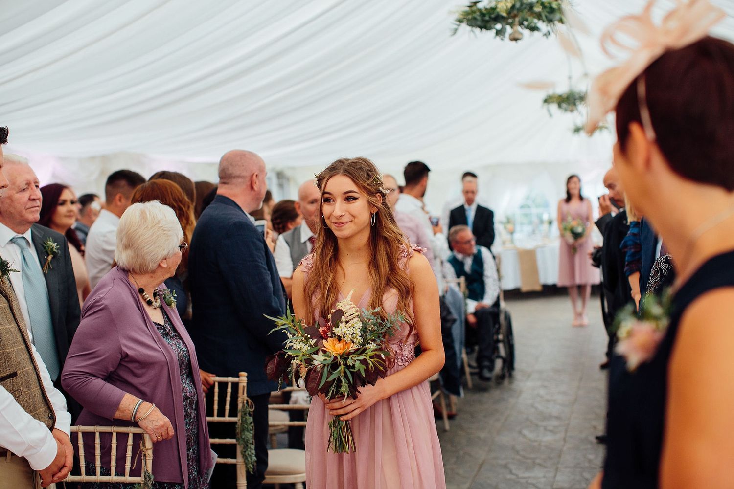 Wedding ceremony scene with guests seated in a white tent decorated with draped fabric.