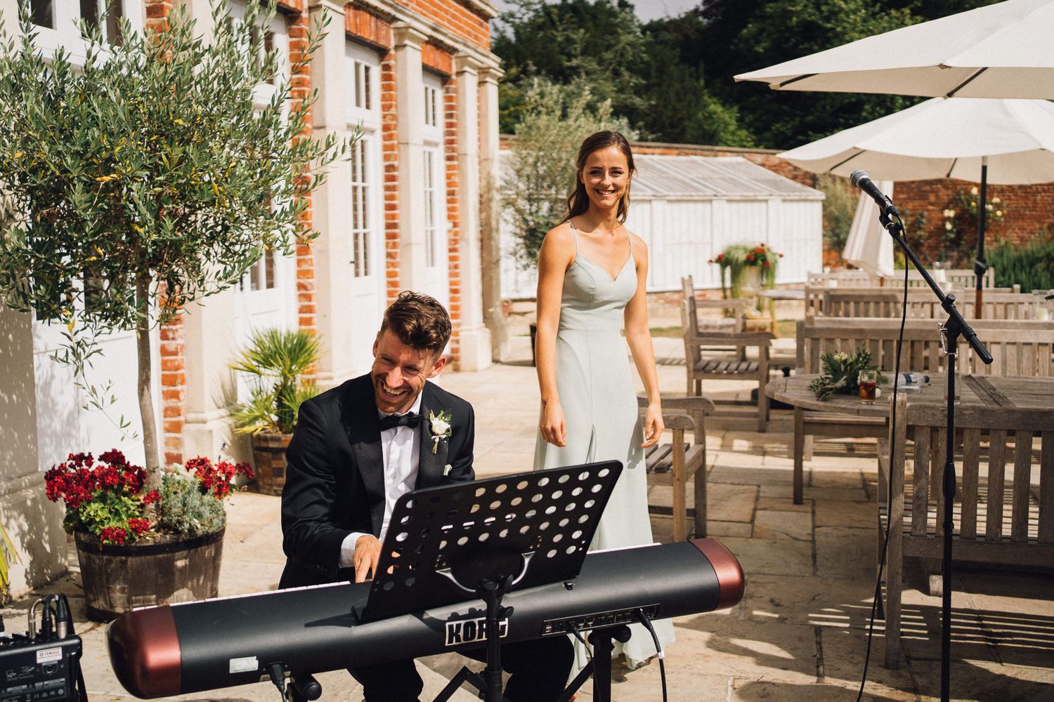 Musicians perform live music at an elegant outdoor wedding reception with brick architecture and white umbrellas.