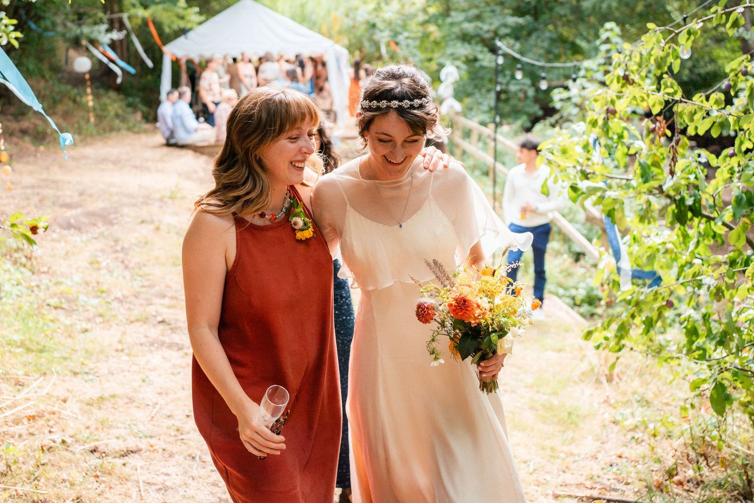 Bride in white gown holding colorful bouquet walks with smiling guest in rust dress at outdoor wedding celebration.