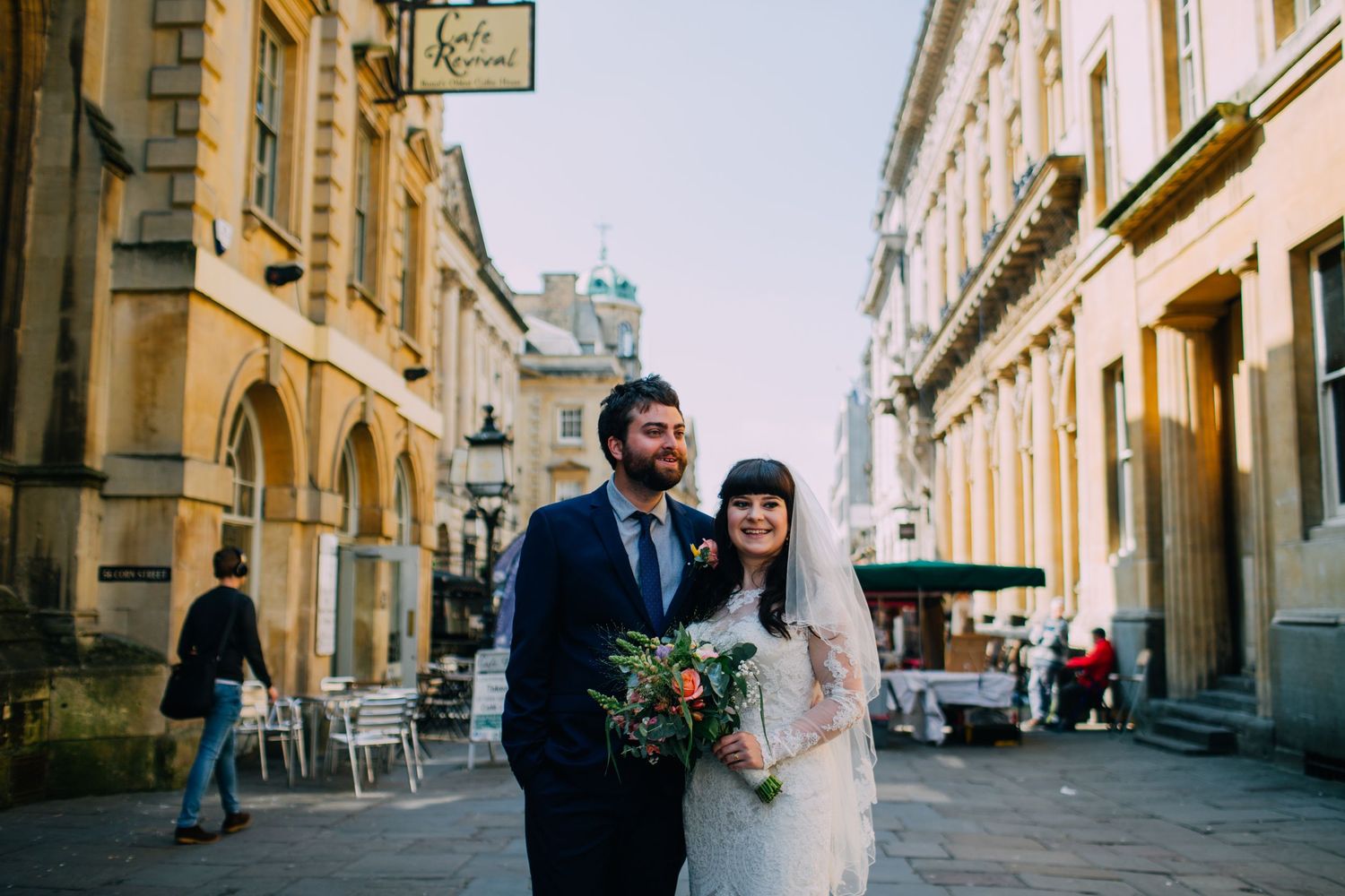 Bride and groom pose on a cobblestone European street with historic architecture and a café in the background.