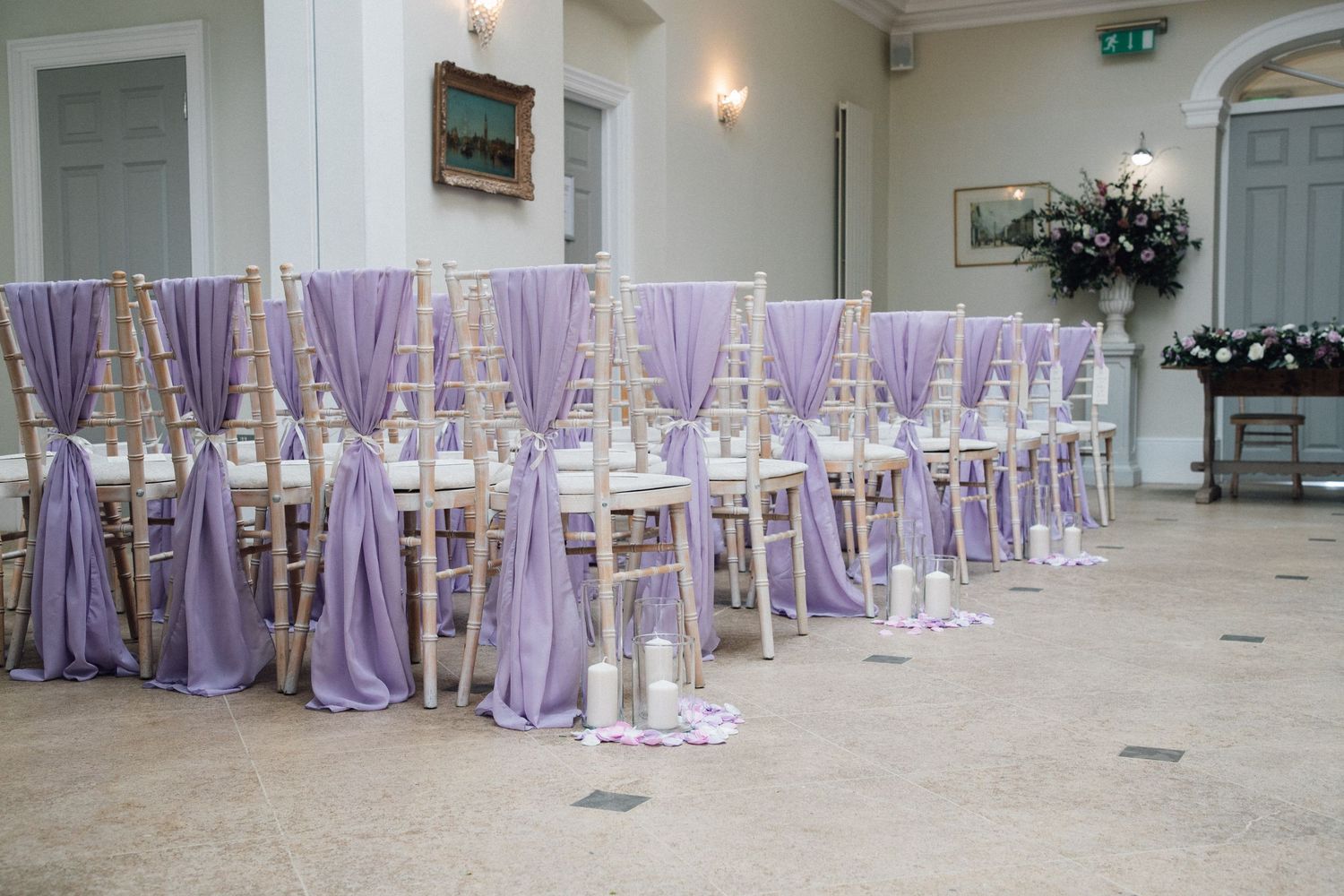 Elegant wedding ceremony chairs adorned with lavender chiffon sashes arranged in rows on a marble floor in a grand venue.