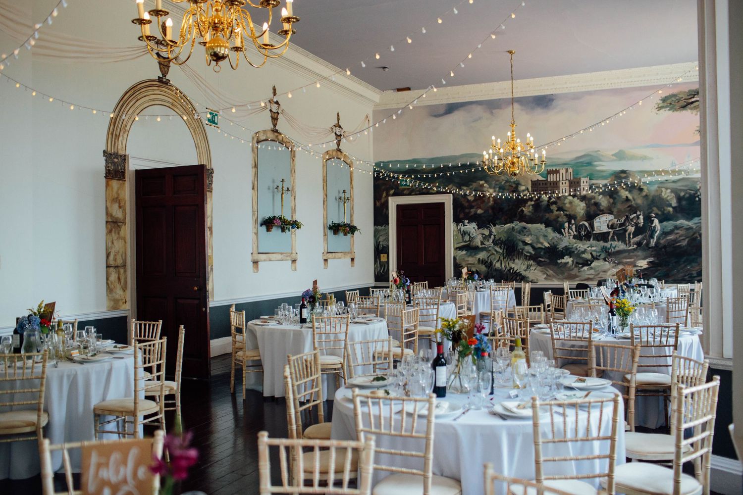 Elegant wedding reception hall with round tables, gold chairs, fairy lights, chandeliers, and a scenic mural on the wall.