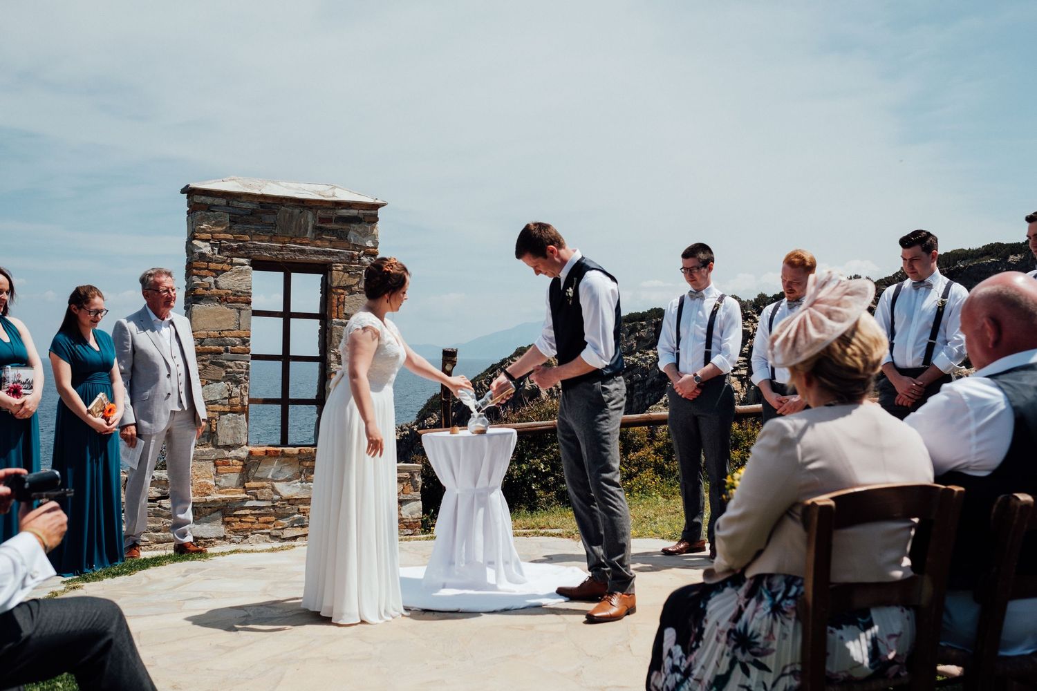 Wedding celebration by the sea with guests mingling on stone patio near water.