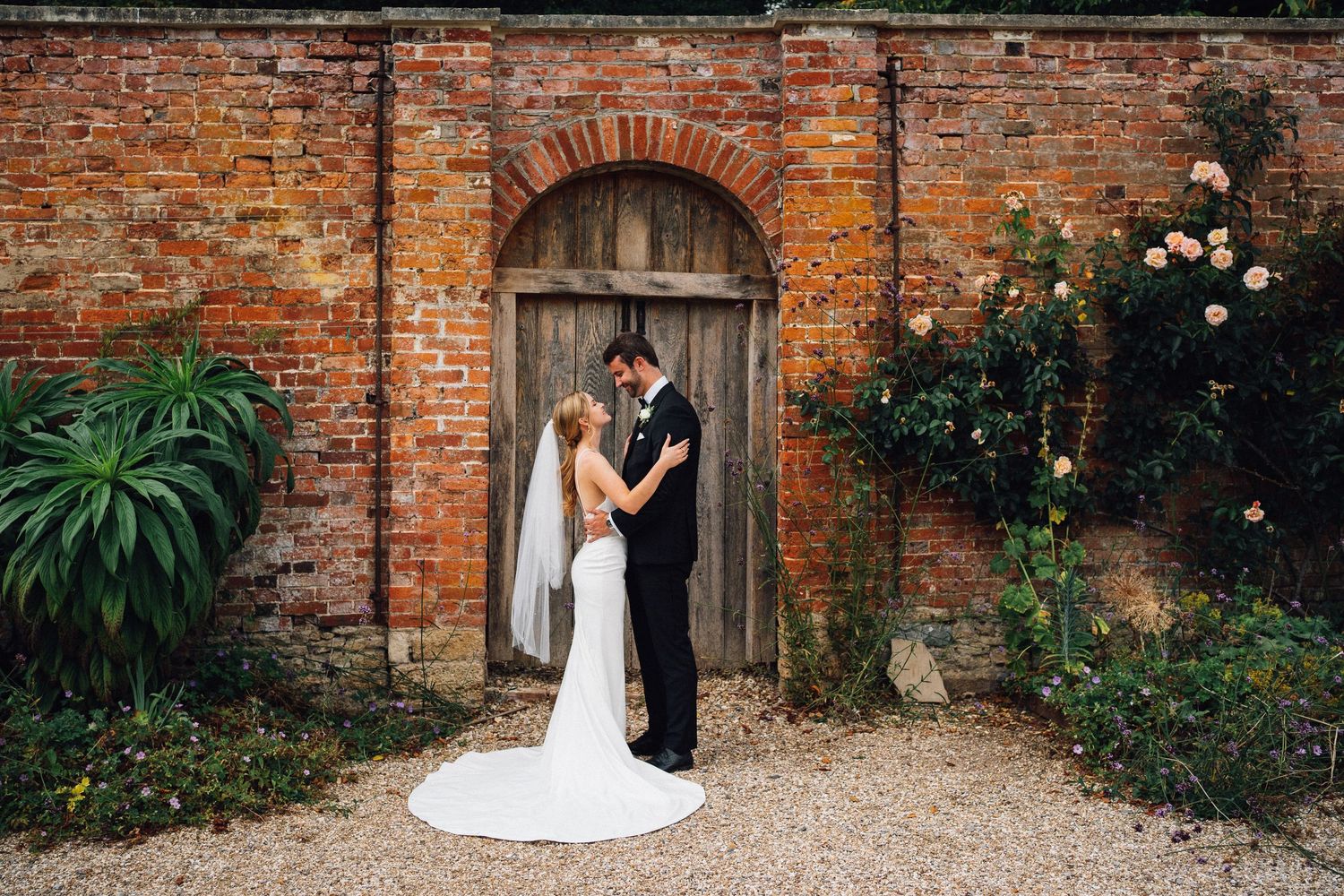 Newlyweds embrace in front of rustic brick archway with climbing roses and greenery.
