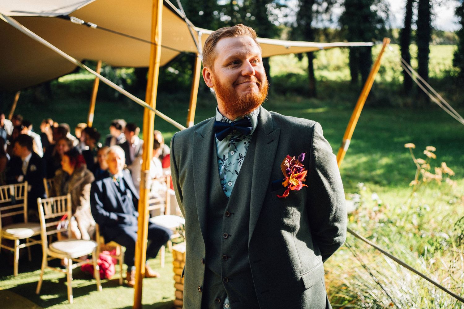 A groom in a dark suit and bow tie smiles outdoors at a tented wedding ceremony with wooden chairs.