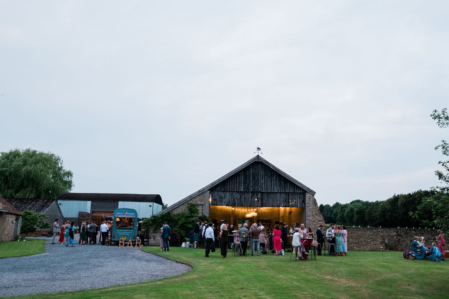 Rural event space with multiple wooden barns and guests mingling on sprawling green grounds.