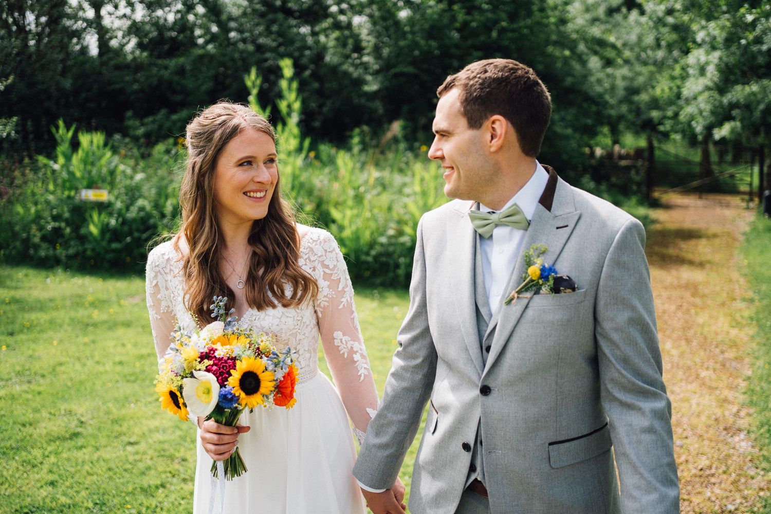 Newlyweds walk hand in hand through a garden path carrying sunflower bouquet.