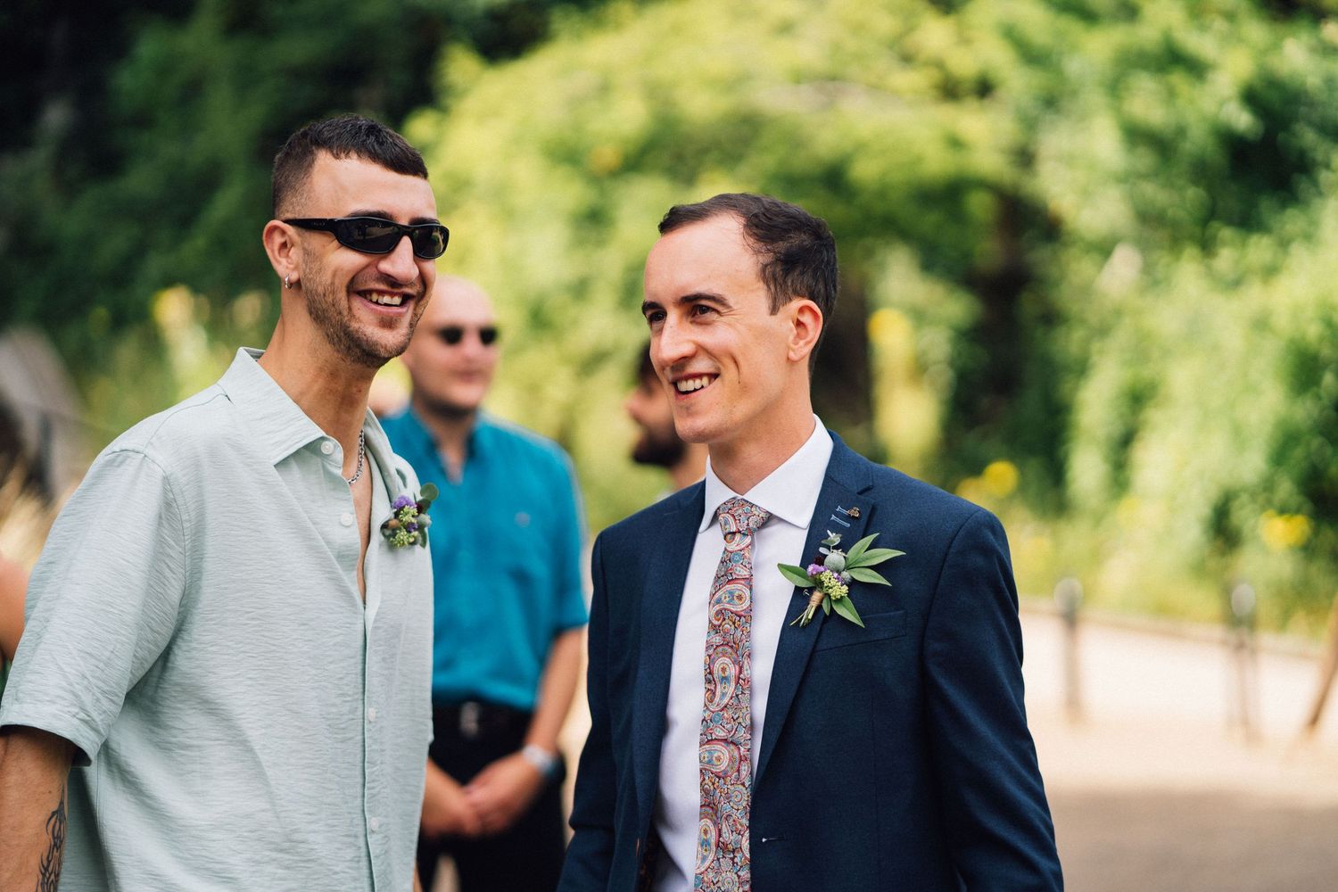 Two people share a joyful moment at an outdoor wedding celebration, wearing sunglasses and floral boutonnieres.