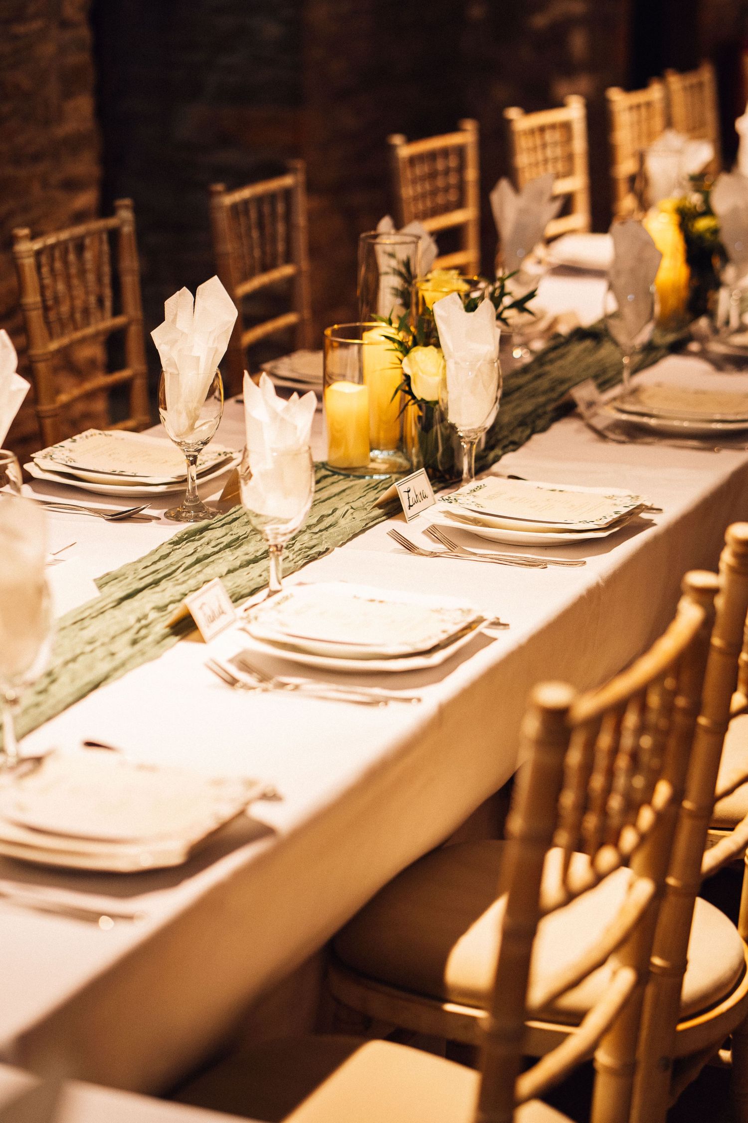 Long dining table with gold chairs and white place settings illuminated by soft candlelight.