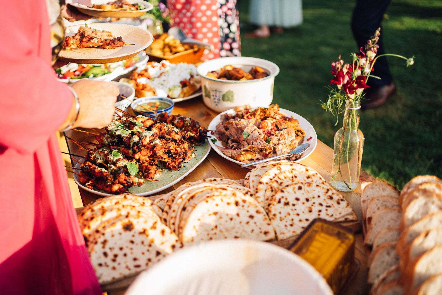 A rustic outdoor wedding reception table decorated with pink lighting and various food dishes.