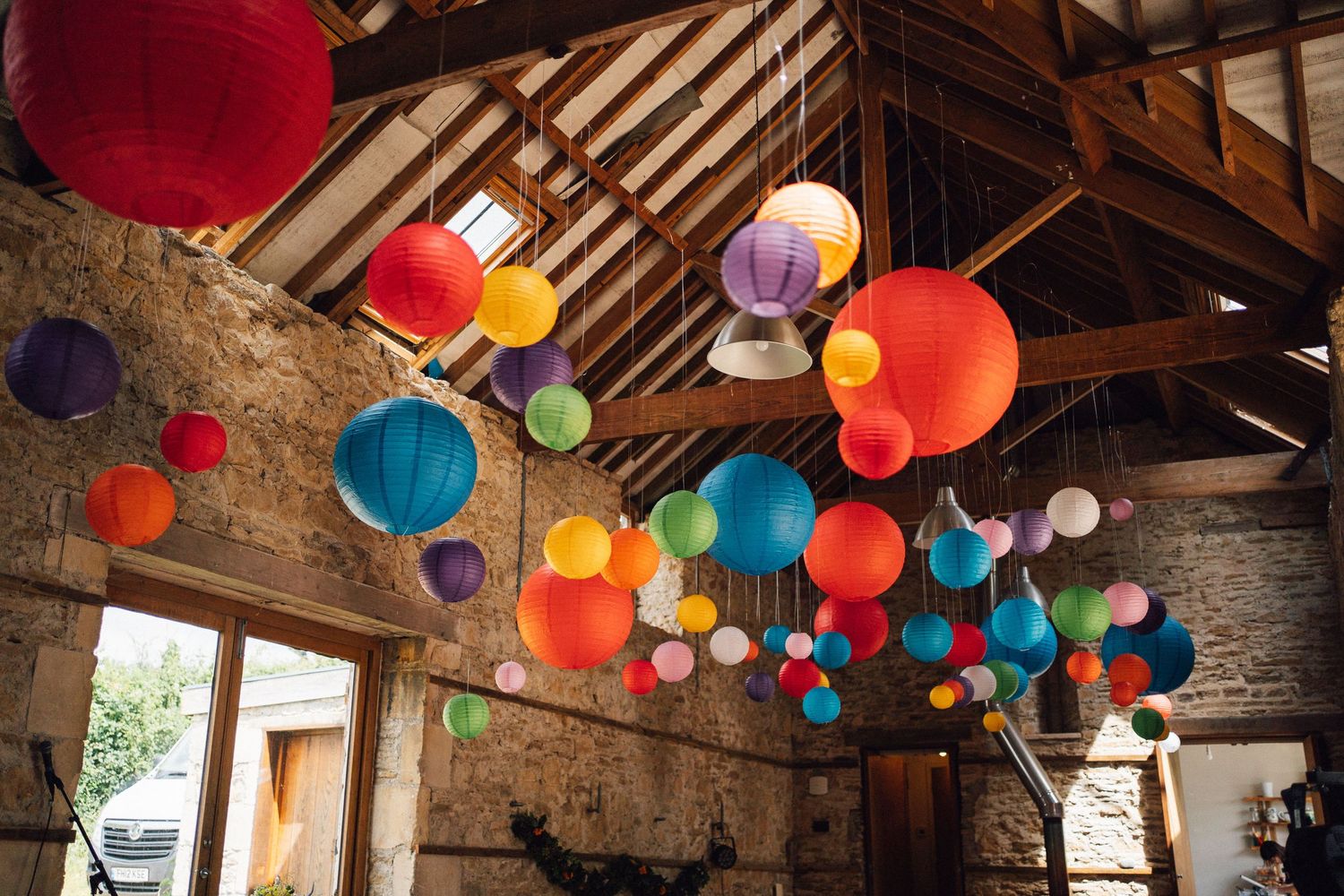 Colorful paper lanterns hanging from rustic barn ceiling beams in red, blue, green, yellow, and purple hues.