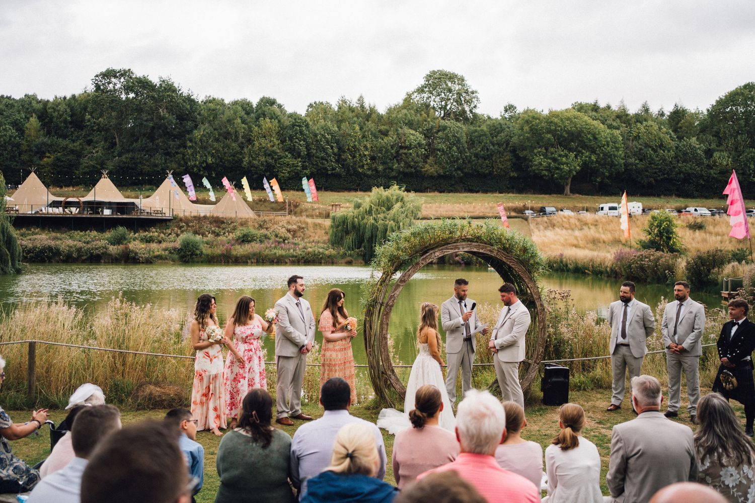 An outdoor wedding ceremony takes place near a lake with teepee tents visible in the background.