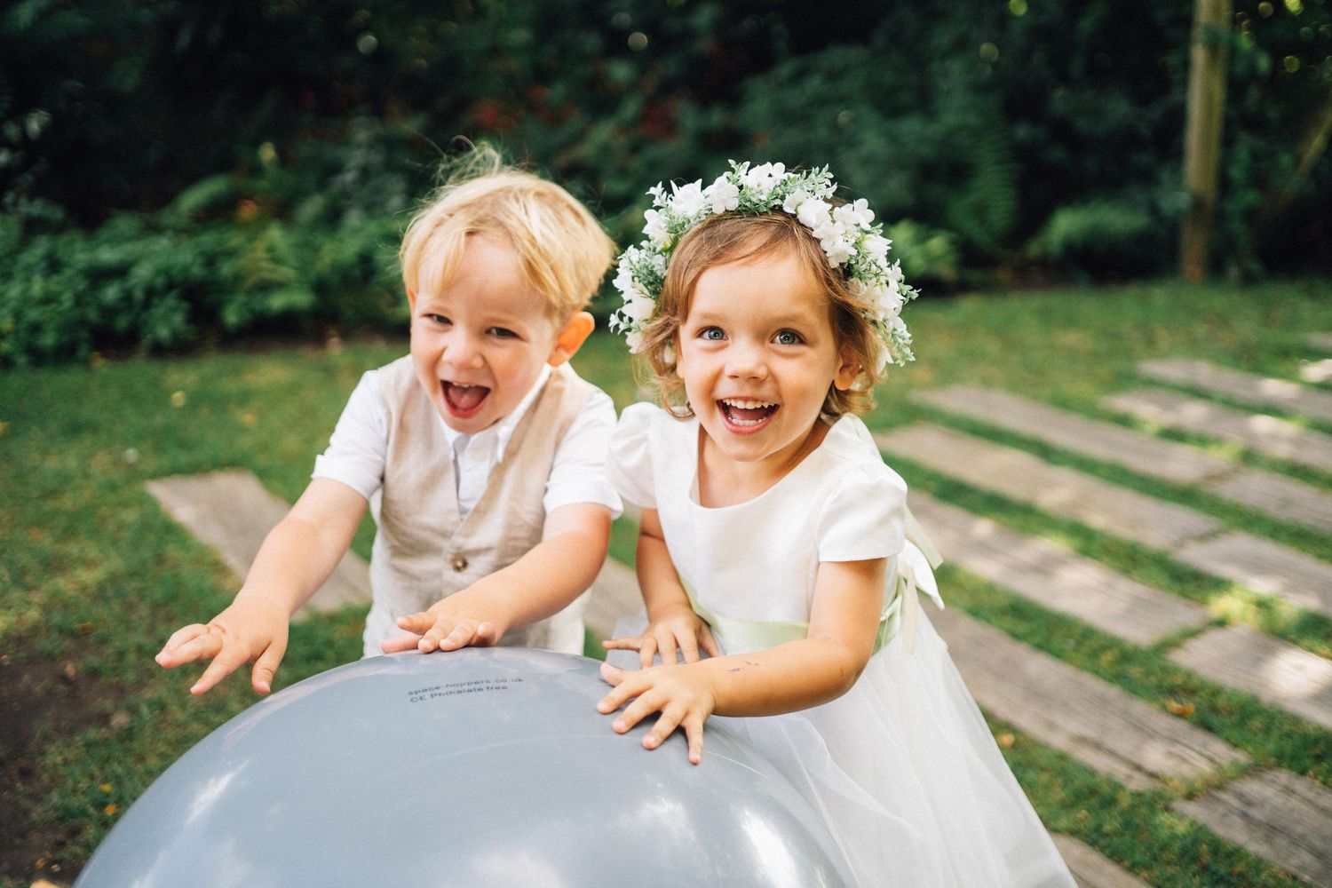 Two joyful children in white wedding attire push a large balloon in a garden setting.