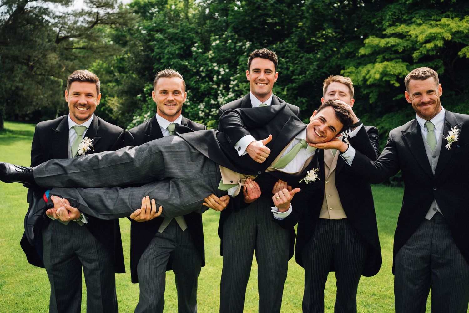 Groomsmen in formal attire strike playful poses on grass during wedding photos.
