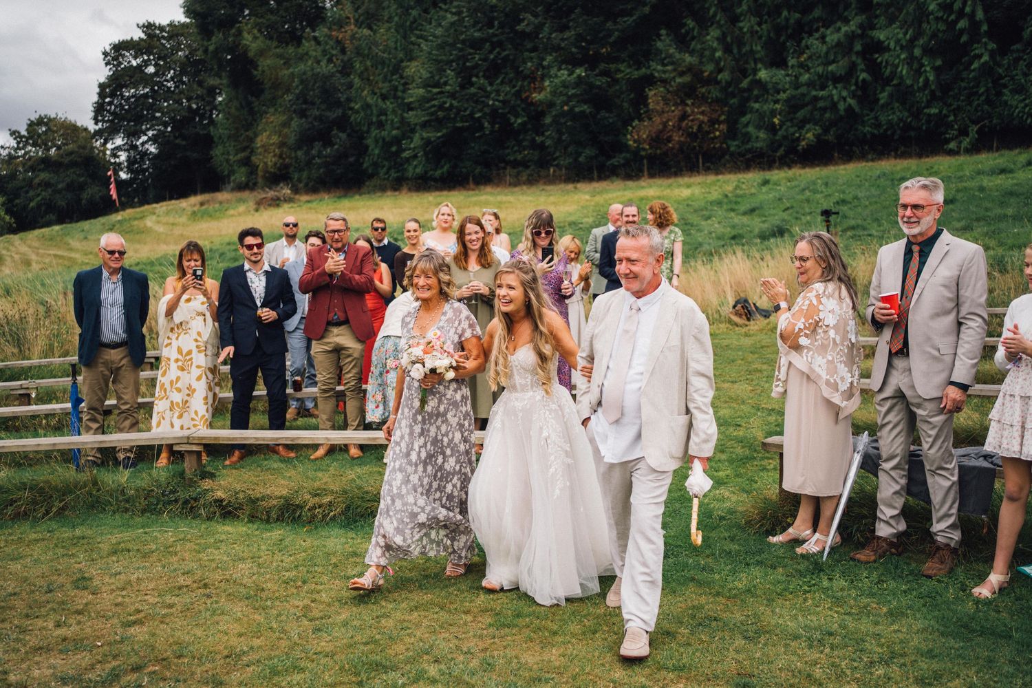 Wedding party and guests walk together through a grassy field during celebration.