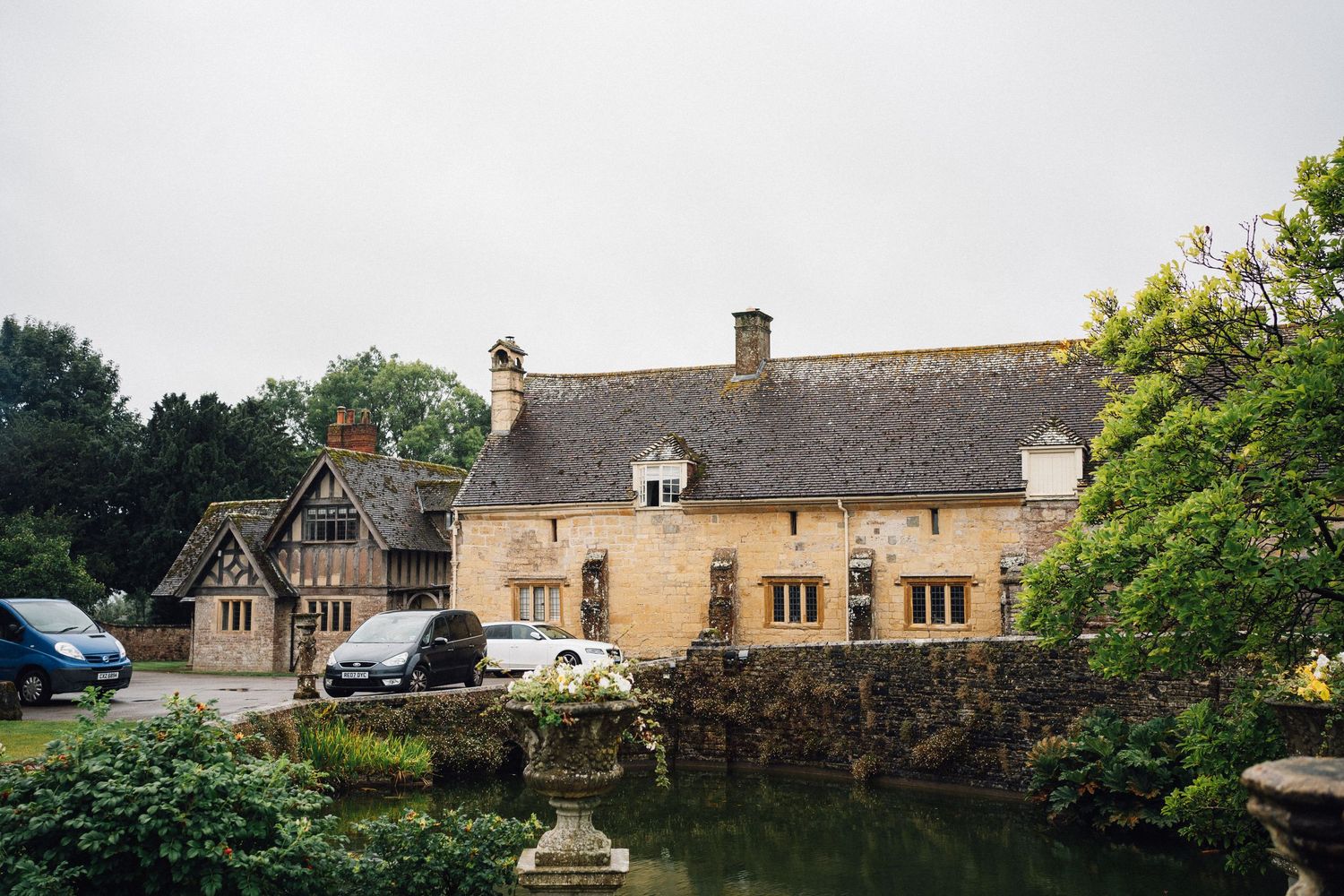 A historic stone building with Tudor-style architecture sits beside a pond on a cloudy day.