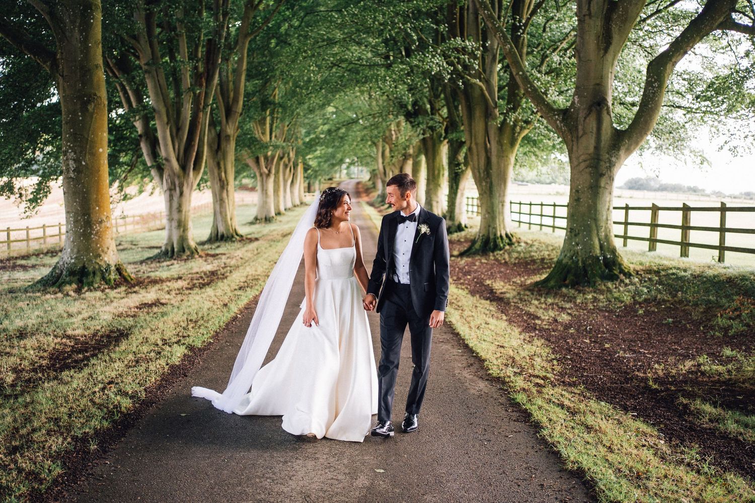 Newlyweds stroll hand in hand down a scenic tree-lined pathway on their wedding day.