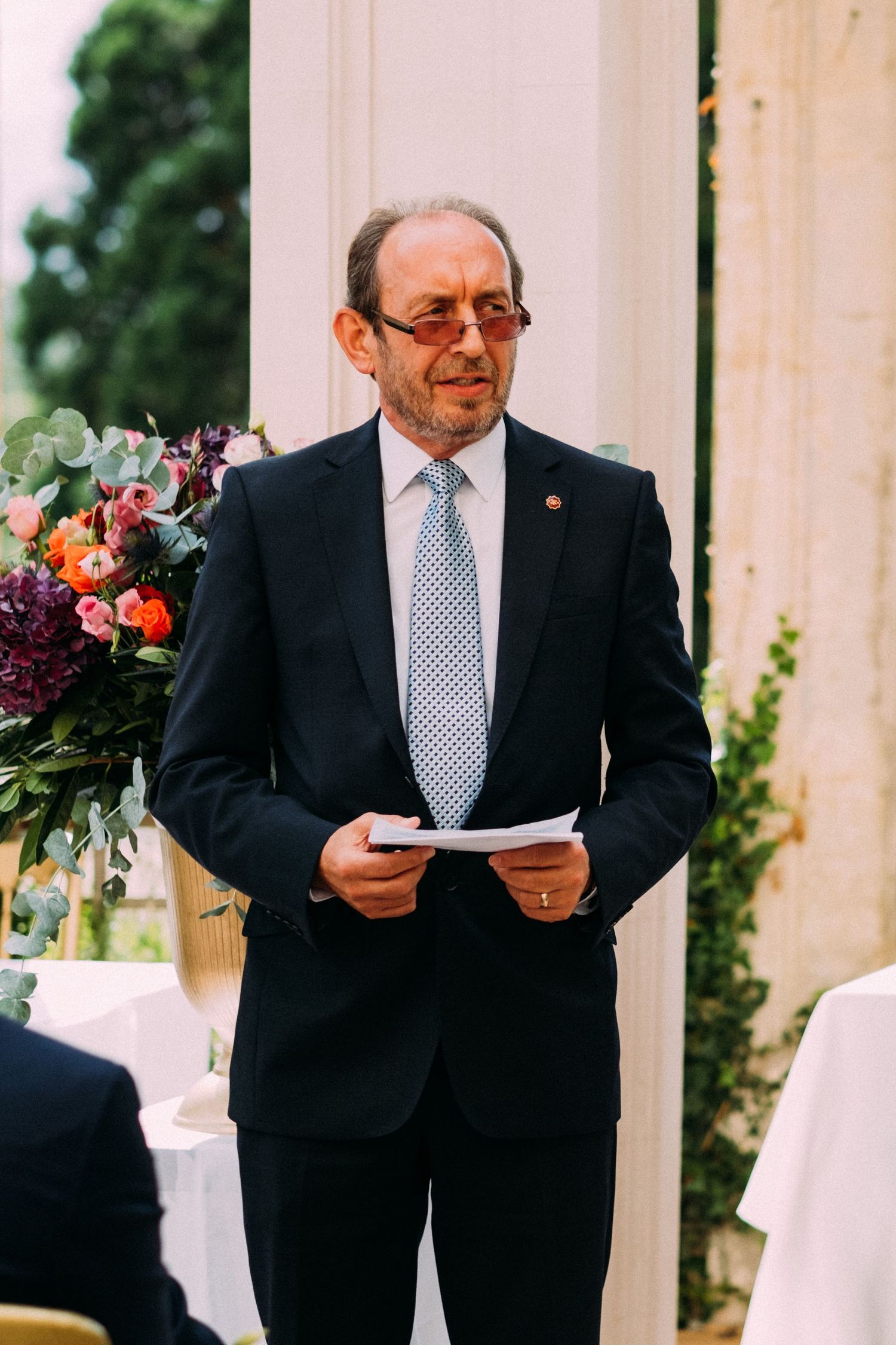 An officiant in a suit and tie reads from papers during an outdoor wedding ceremony.