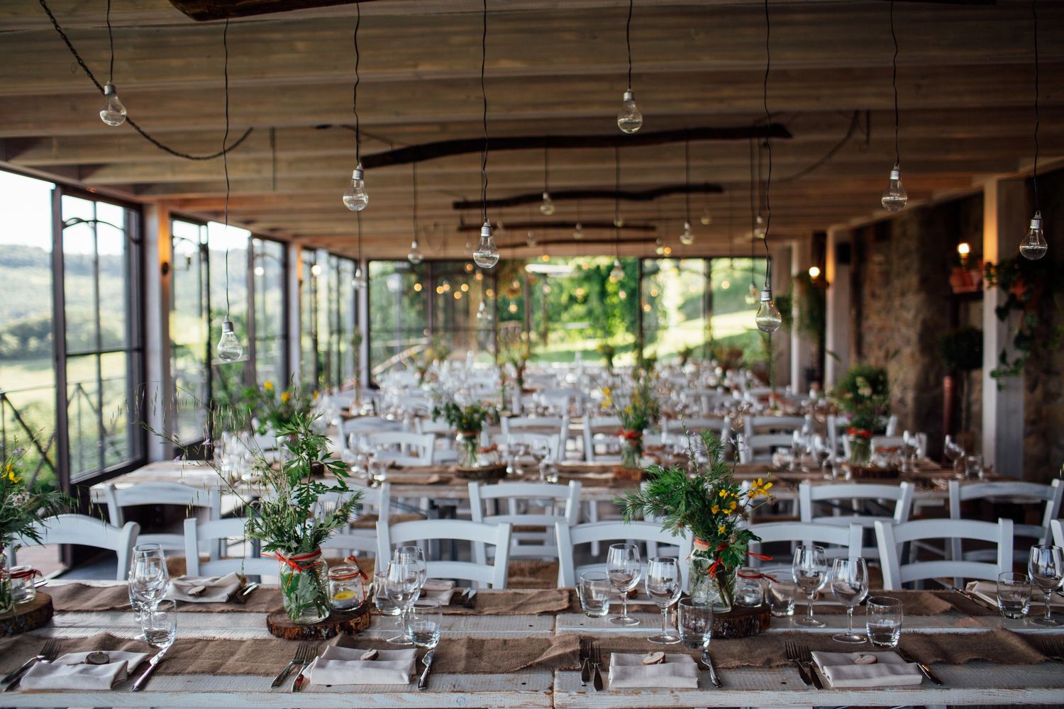 Wedding reception hall decorated with white tables and chairs under exposed wooden beams and string lights.