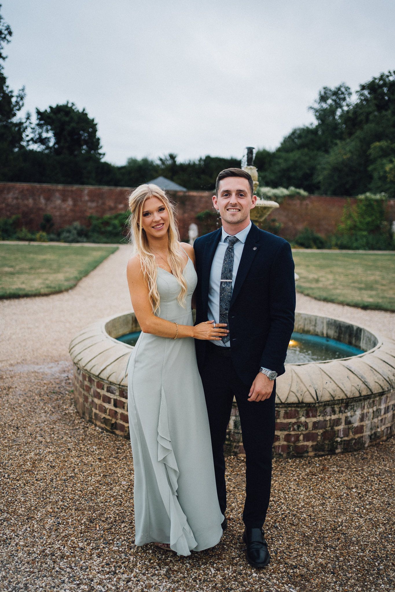 A couple poses together by a brick fountain during an elegant outdoor wedding celebration.