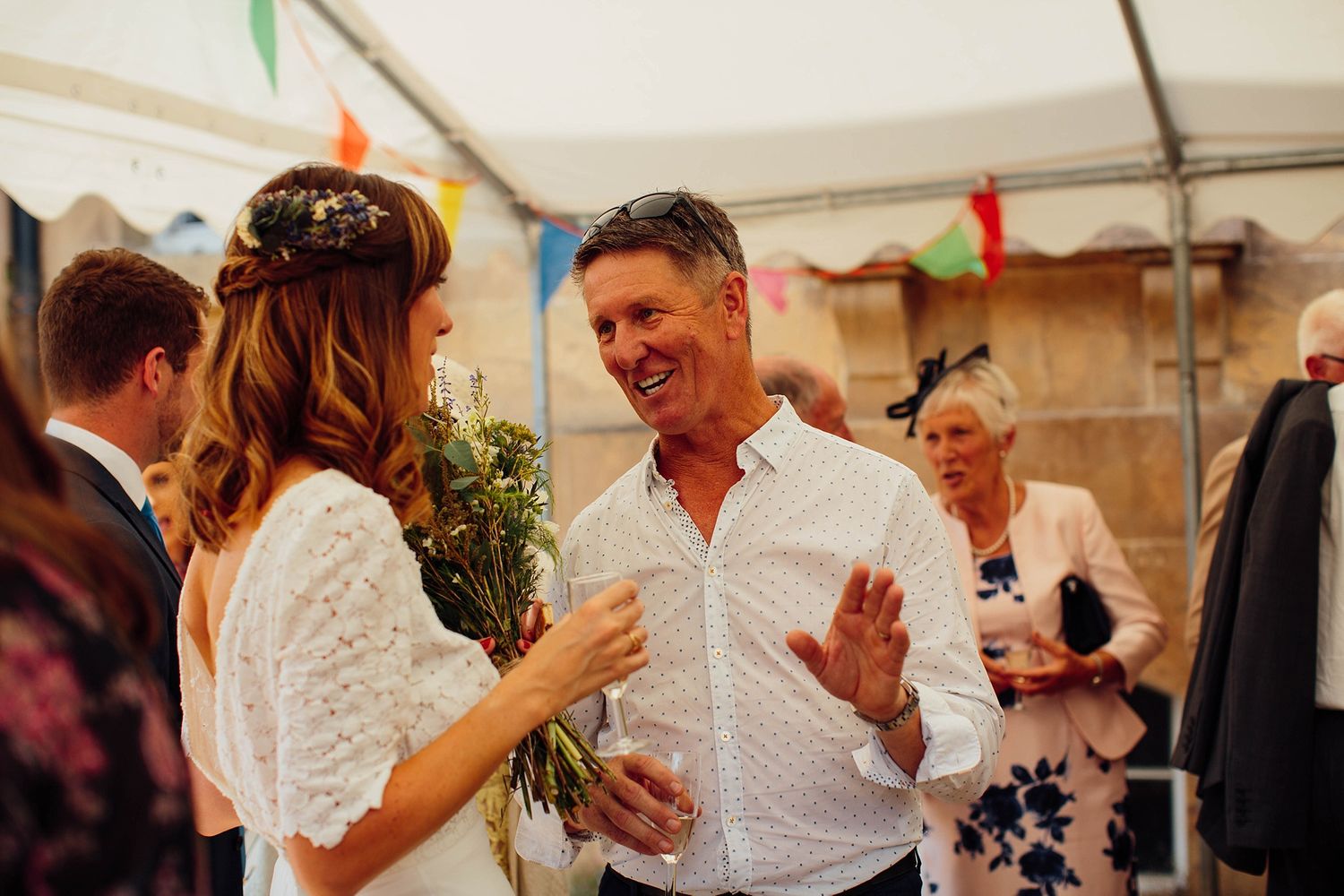 Wedding guests laughing and socializing under festive tent with bunting decorations.