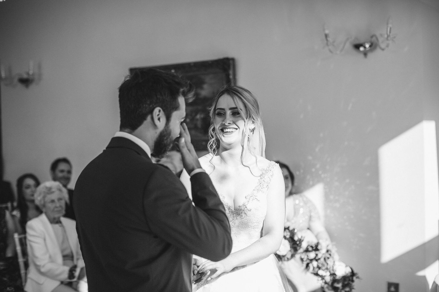 Black and white photo captures an intimate wedding ceremony moment in a sunlit room.