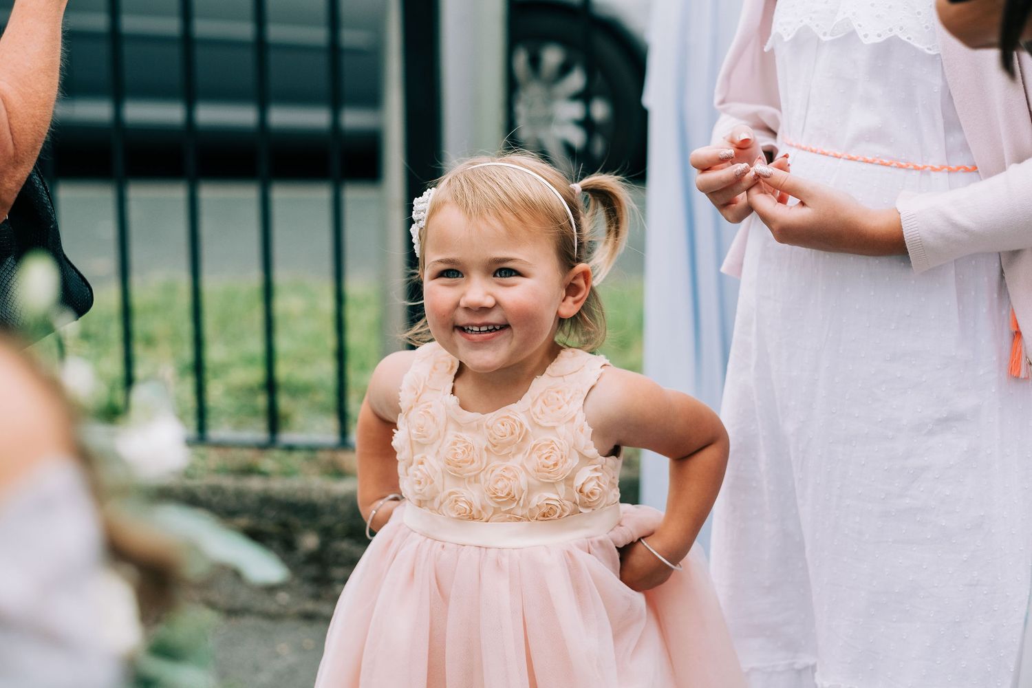 Young flower girl in a peach dress smiling brightly during outdoor wedding festivities.