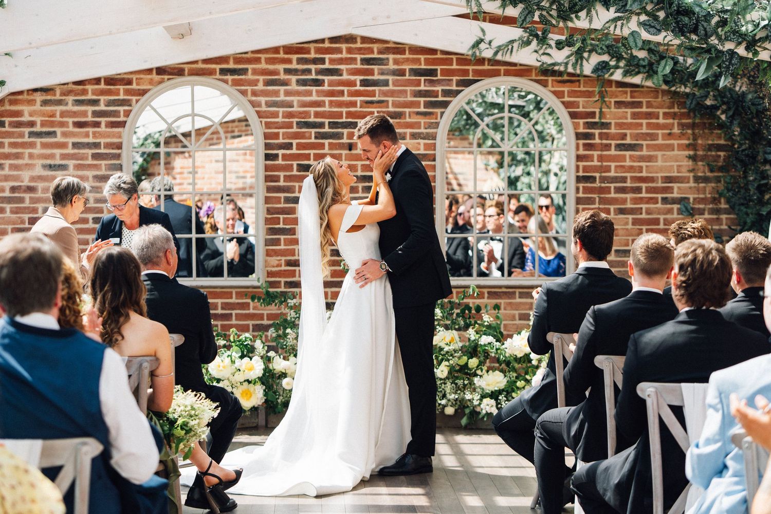 Wedding ceremony taking place in an elegant brick venue with arched windows and guests seated in rows.