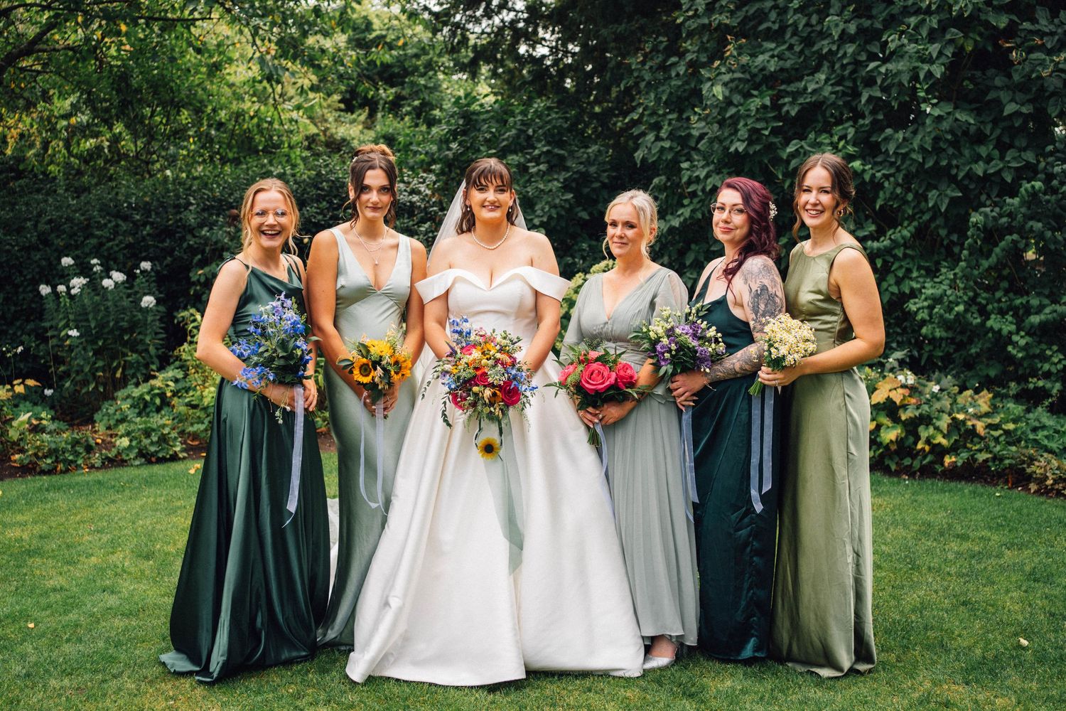 Wedding party poses on lawn with bridesmaids in sage green dresses holding colorful bouquets.