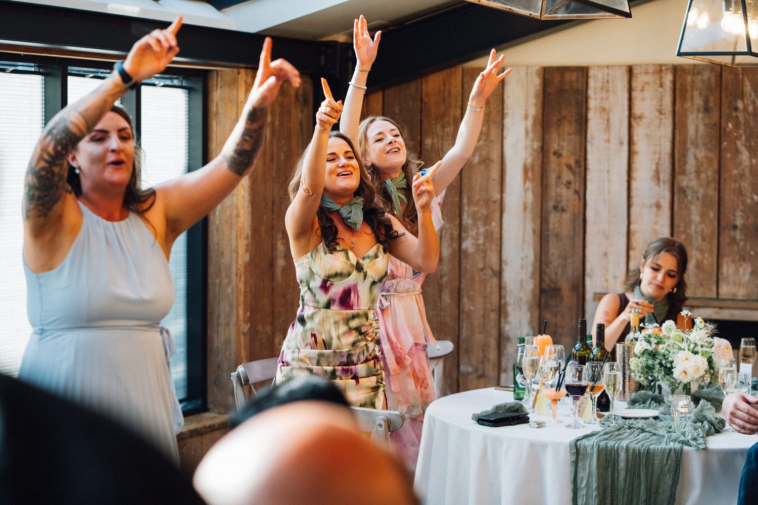 Wedding reception guests raising glasses in celebration at rustic wooden venue with string lights.