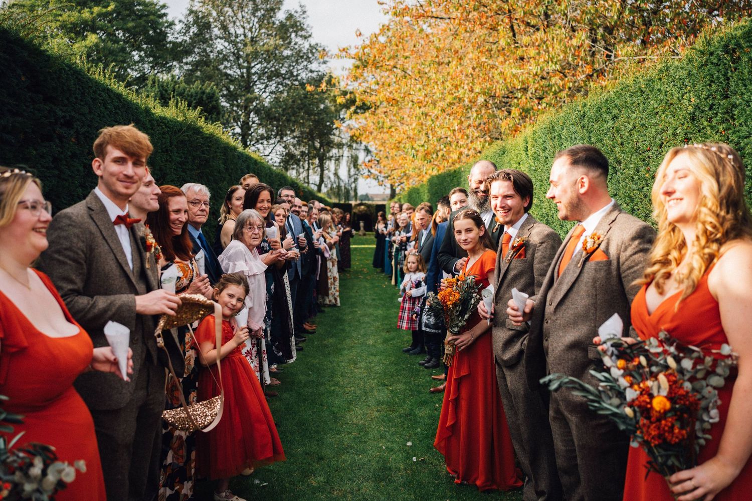 Wedding party guests mingle outdoors on a grassy hill during autumn with golden trees in the background.