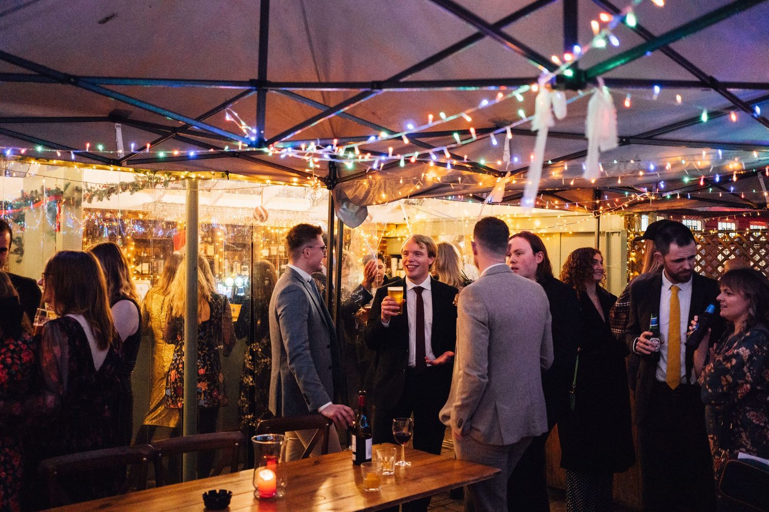 String lights illuminate a lively evening wedding reception under a white tent with guests mingling and dancing.