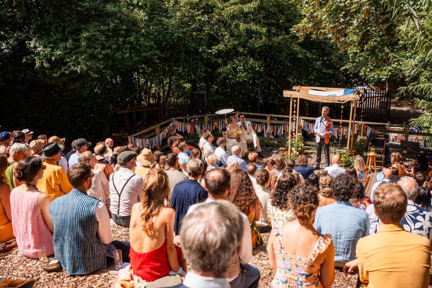 Outdoor audience watches a performer on a wooden stage surrounded by lush green trees on a sunny day.