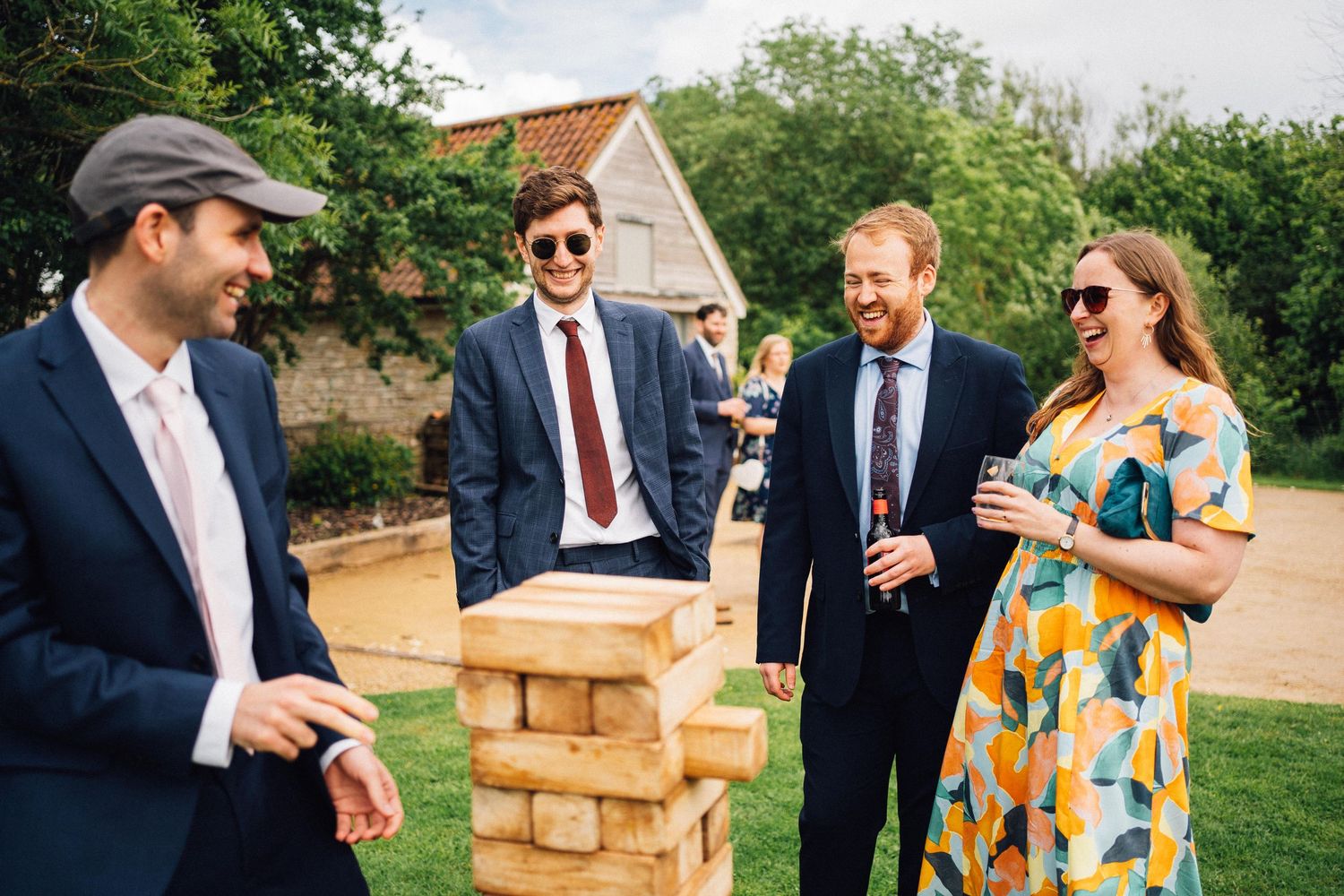 Wedding guests play giant Jenga outdoors at a rustic venue during a summer celebration.