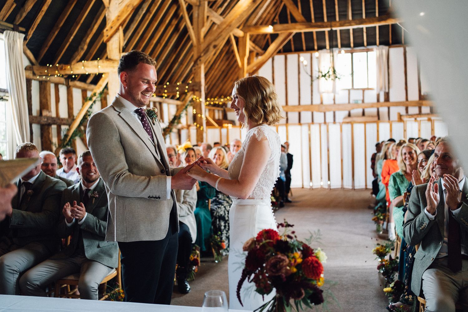 A couple exchanges vows during a rustic barn wedding ceremony with exposed wooden beams and guests watching.