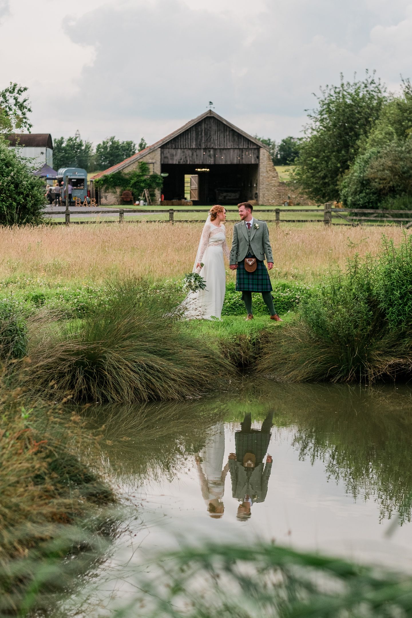 Two people stand at the edge of a serene pond surrounded by tall grass and rustic farm buildings in the background.