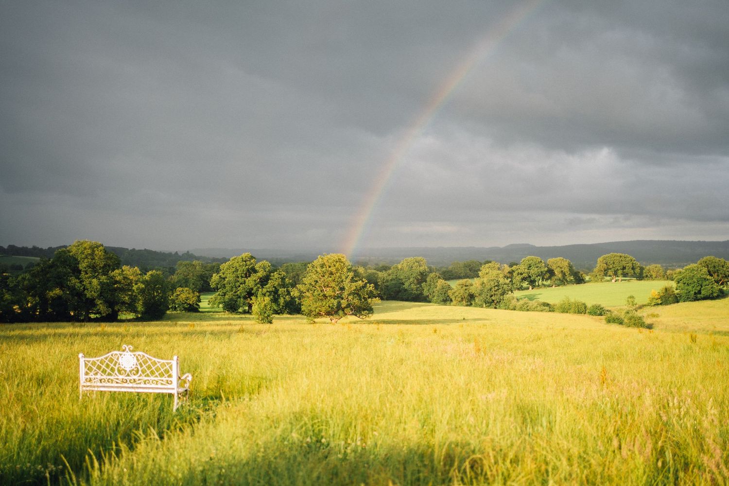 Panoramic view of rolling green hills and fields under dramatic stormy skies with a rainbow.