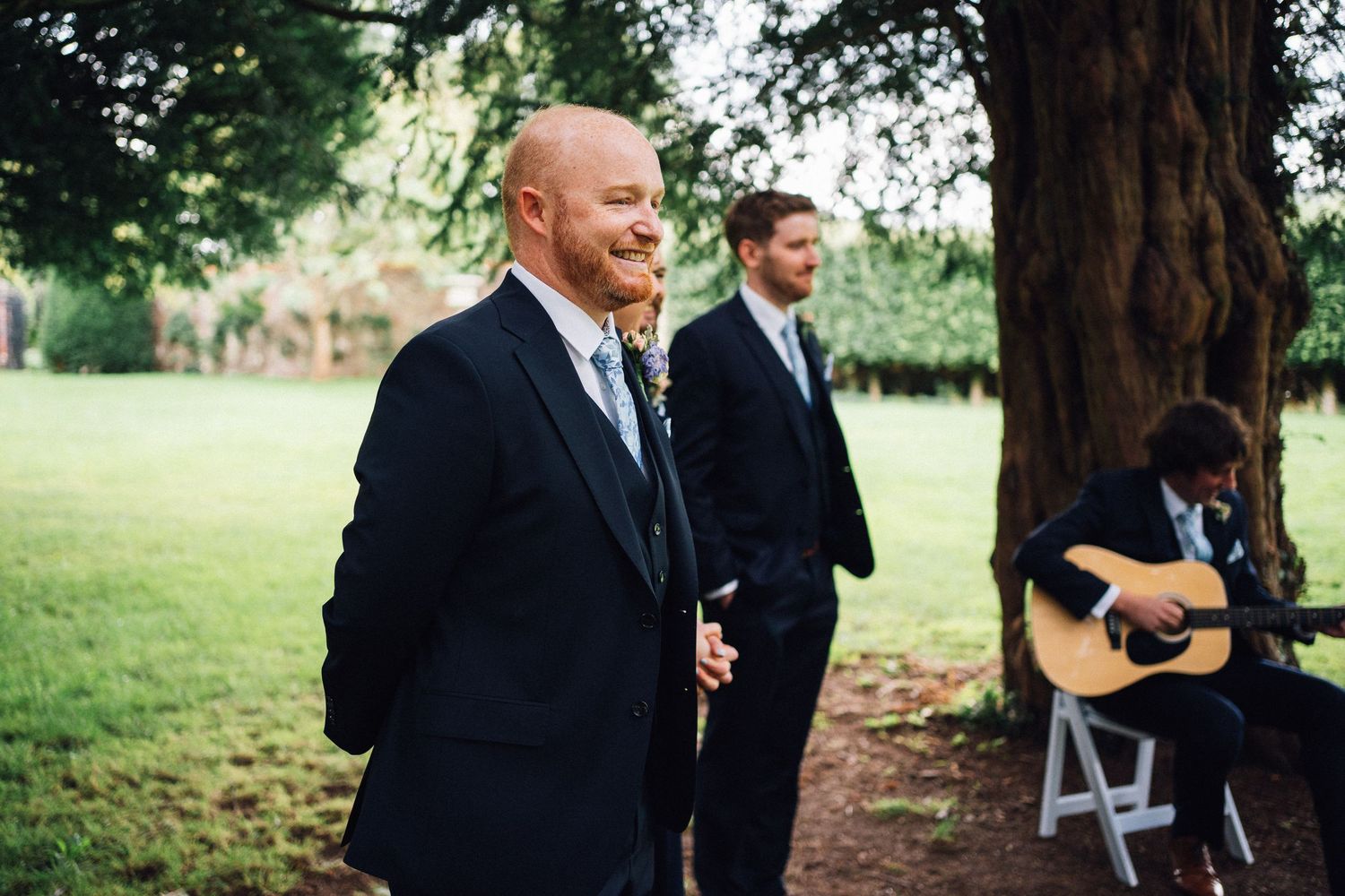 Outdoor wedding ceremony under tall trees with white chairs and floral arrangements.