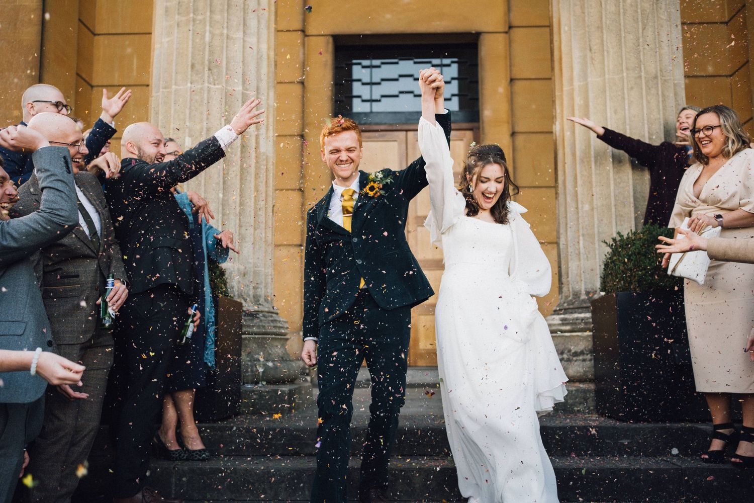 Wedding guests throw confetti outside a historic stone building with columns.