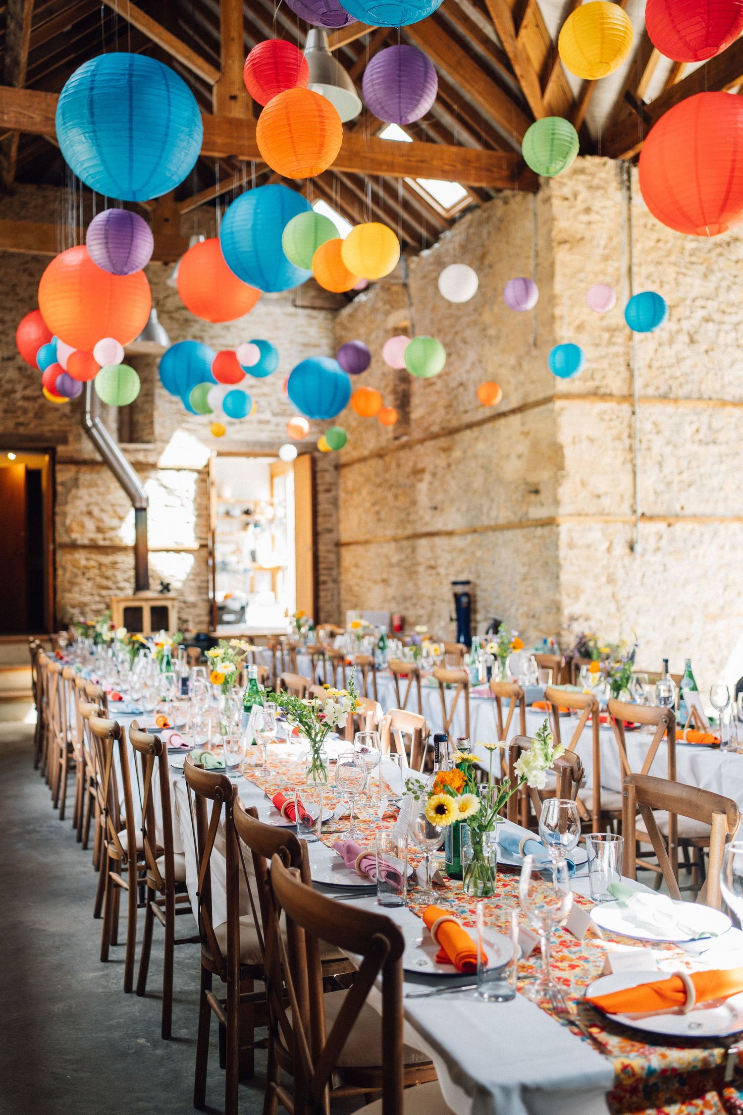 Colorful paper lanterns decorate rustic reception venue with long wooden tables and place settings.