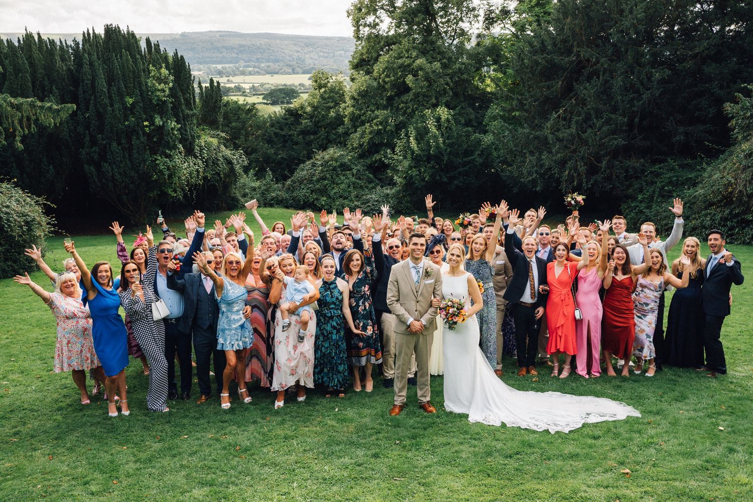 Large group of wedding guests gathered outdoors on a grassy lawn for celebratory photos.