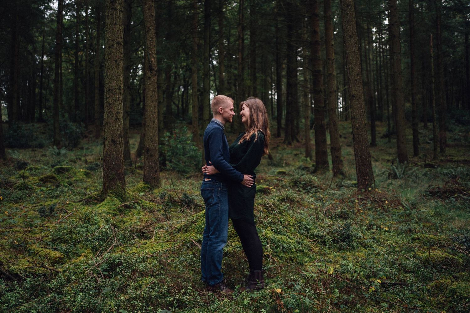 Two people sharing an intimate moment among tall trees in a forest clearing.