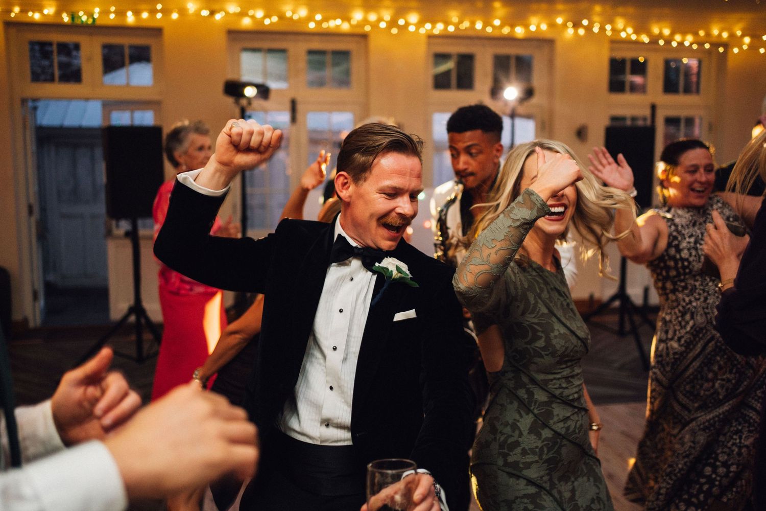 Groom in tuxedo dancing joyfully with guests at elegant wedding reception with string lights.